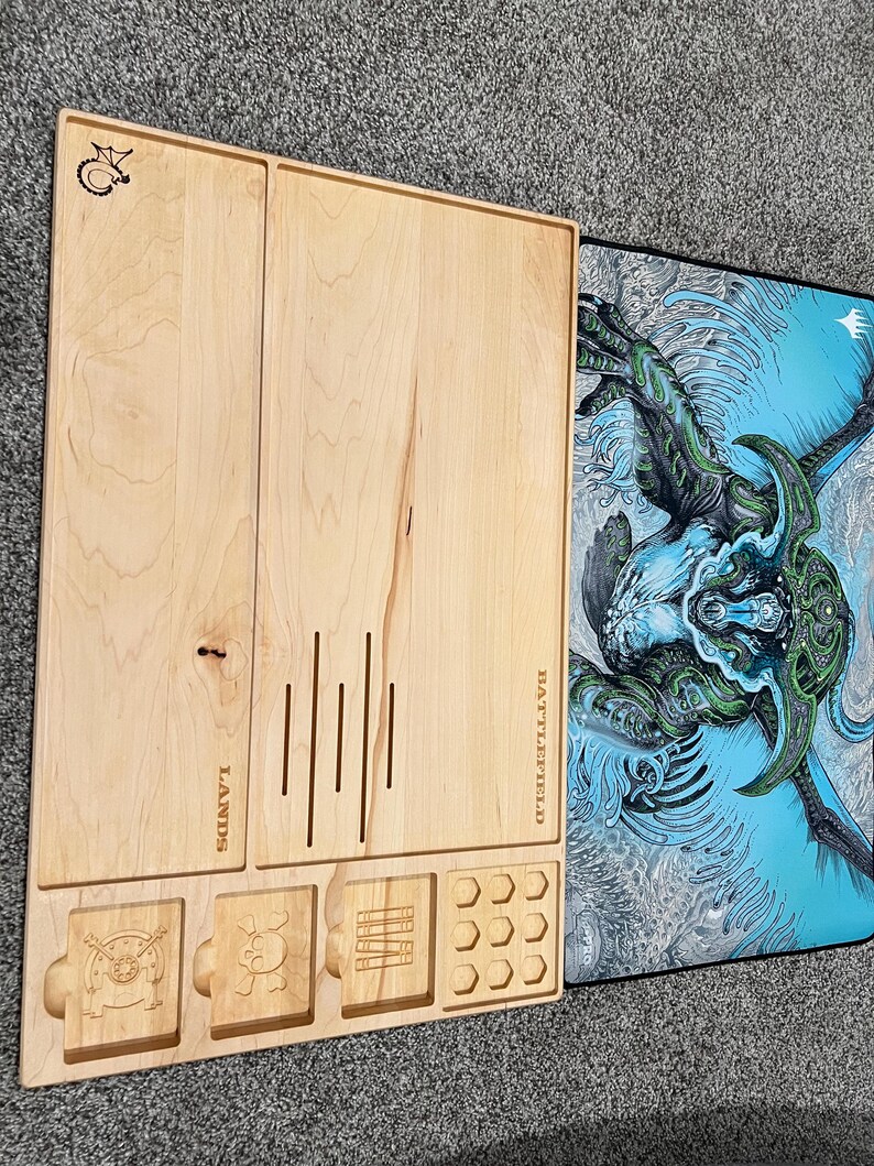 May include: A light-colored wooden game board with compartments and etched designs. The board has slots for cards, dice, and tokens, with the word "LANDS" and "BATTLEFIELD" etched on it. A blue and green graphic mat is next to the board.