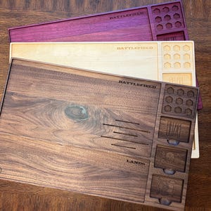 May include: Three wooden game trays in shades of brown, cream, and purple. Each tray has compartments and the word "BATTLEFIELD" engraved on it. The bottom tray also has the word "LANDS" engraved.
