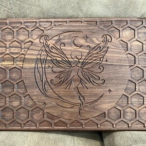 May include: A rectangular wooden serving board with a carved design. The board features a central image of a fairy on a crescent moon, surrounded by stars. The border is decorated with a honeycomb pattern. The wood has a dark brown finish.
