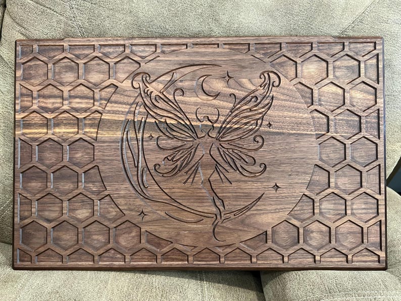 May include: A rectangular wooden serving board with a carved design. The board features a central image of a fairy on a crescent moon, surrounded by stars. The border is decorated with a honeycomb pattern. The wood has a dark brown finish.