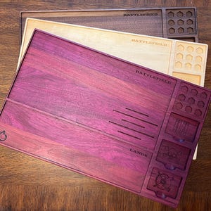 May include: Three wooden game trays in shades of purple, natural, and brown. The purple tray has the word "BATTLEFIELD" and "LANDS" engraved on it, along with various compartments and slots. The other trays also have the word "BATTLEFIELD" engraved.
