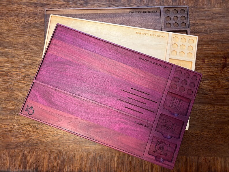 May include: Three wooden game trays in shades of purple, natural, and brown. The purple tray has the word "BATTLEFIELD" and "LANDS" engraved on it, along with various compartments and slots. The other trays also have the word "BATTLEFIELD" engraved.