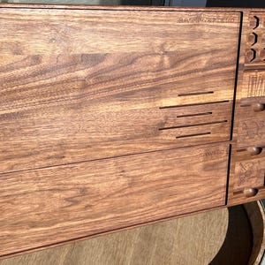 May include: A wooden tabletop organizer with multiple compartments and drawers. The organizer is made of dark brown wood and features the text "BATTLEFIELD" and "LANDS" etched into the surface. It includes various storage slots and engraved designs.