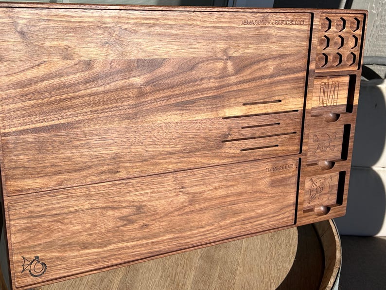 May include: A wooden tabletop organizer with multiple compartments and drawers. The organizer is made of dark brown wood and features the text "BATTLEFIELD" and "LANDS" etched into the surface. It includes various storage slots and engraved designs.