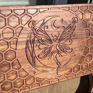May include: A rectangular wooden tray with a carved design. The tray features a fairy with large wings, a crescent moon, and stars. The border has a honeycomb pattern. The wood has a warm, brown tone.