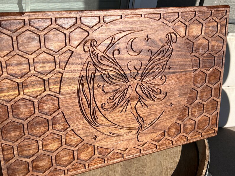 May include: A rectangular wooden tray with a carved design. The tray features a fairy with large wings, a crescent moon, and stars. The border has a honeycomb pattern. The wood has a warm, brown tone.