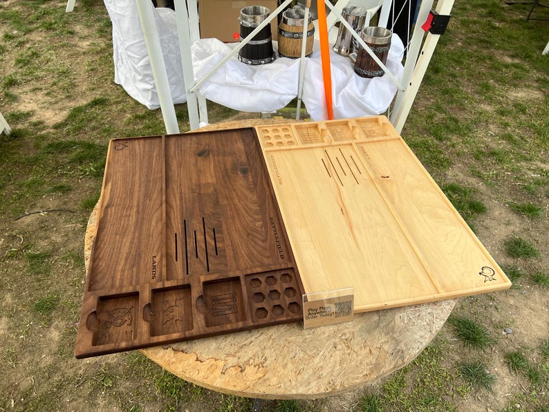 May include: Two wooden game boards, one dark brown and one light brown, are displayed. The dark brown board has carved slots and images, while the light brown board has etched lines. Both boards are rectangular and sit on a round wooden table.