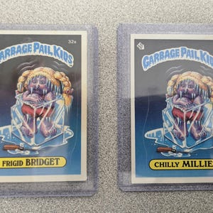 May include: Two Garbage Pail Kids trading cards in protective sleeves. One card features "Frigid Bridget" and the other "Chilly Millie." Both cards depict a cartoon character encased in ice, with the Garbage Pail Kids logo at the top.