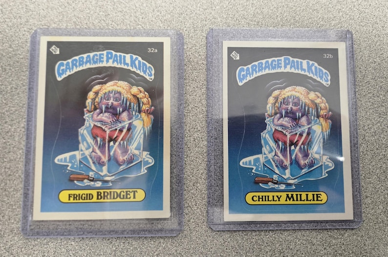 May include: Two Garbage Pail Kids trading cards in protective sleeves. One card features "Frigid Bridget" and the other "Chilly Millie." Both cards depict a cartoon character encased in ice, with the Garbage Pail Kids logo at the top.