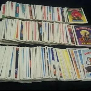 Könnte beinhalten: Eine Sammlung von Vintage-Sammelkarten der Garbage Pail Kids, in Stapeln angeordnet. Die Karten zeigen farbenfrohe Illustrationen und humorvolle Charaktere. Einige Karten sind sichtbar, darunter "Jay Decay" und "Reuben Cube".