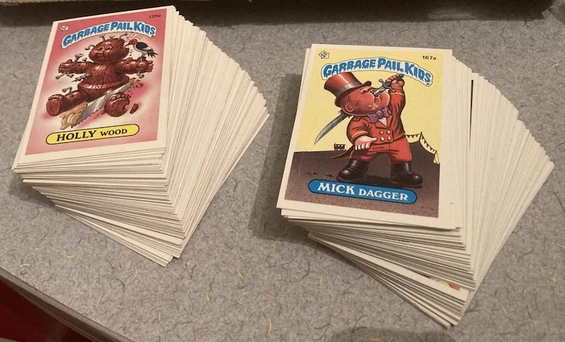 May include: Stacks of vintage Garbage Pail Kids trading cards. One stack features a card with a brown character and the text "HOLLY WOOD". The other stack shows a card with a character in a red suit and the text "MICK DAGGER".