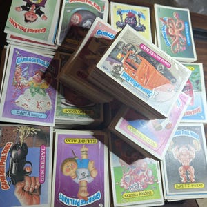 Può includere: Una collezione di figurine vintage Garbage Pail Kids con vari personaggi illustrati con nomi come Dana Druff, Gun Beryl, Elliot Mess, Soggy Oggie, Garbage Mouth Gilbert, Turned-On Ron, Bazooka Joanne e Brett Sweat.