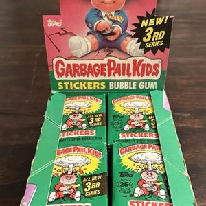 May include: A Garbage Pail Kids sticker bubble gum box set. The box features a cartoon character with a mushroom cloud background. The box set includes multiple packs of stickers and bubble gum. The box is green, red, and yellow.