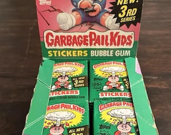 May include: A Garbage Pail Kids sticker bubble gum box set. The box features a cartoon character with a mushroom cloud background. The box set includes multiple packs of stickers and bubble gum. The box is green, red, and yellow.
