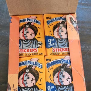 May include: A cardboard box, open to reveal four packs of Garbage Pail Kids stickers. The packs are orange, featuring cartoon illustrations and text stating "9th Series" and "Stickers". Each pack contains 5 stickers and 1 stick of bubble gum.