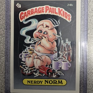 May include: A Garbage Pail Kids trading card featuring "Nerdy Norm." The illustration depicts a baby with multiple cigarettes in its mouth and hands, surrounded by cigarettes, coffee cups, and bottles. The card is in a protective sleeve.