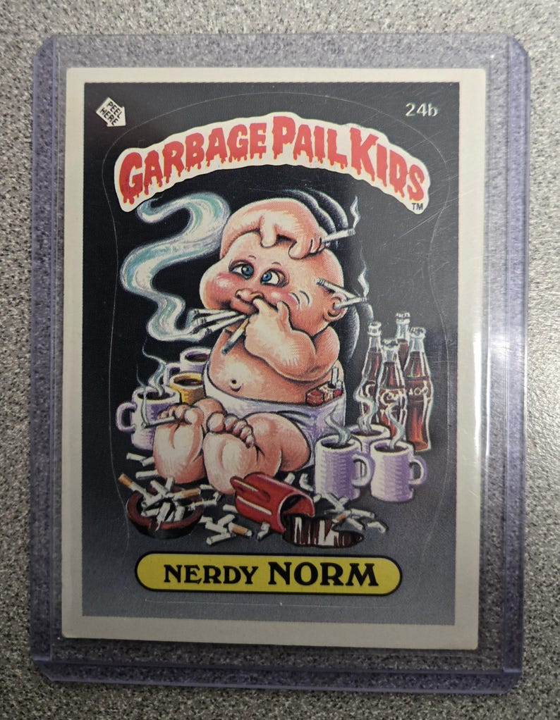 May include: A Garbage Pail Kids trading card featuring "Nerdy Norm." The illustration depicts a baby with multiple cigarettes in its mouth and hands, surrounded by cigarettes, coffee cups, and bottles. The card is in a protective sleeve.