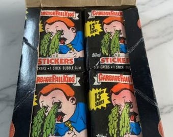 May include: A box of Garbage Pail Kids sticker packs. Each pack features a cartoon character with green vomit and the text "STICKERS". The box is black with orange accents and the text "13th Series".