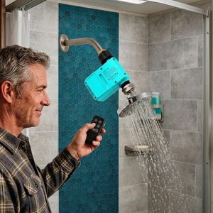Può includere: Un soffione doccia cromato con acqua che spruzza verso il basso. Un dispositivo turchese è collegato al soffione. Una persona tiene in mano un telecomando nero. La doccia è in un bagno con piastrelle grigie e turchese.