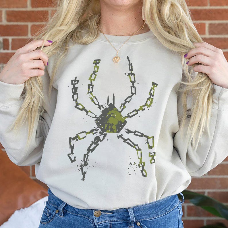 May include: A beige sweatshirt featuring a green and black spider design. The spider graphic is made of chain links and has a camouflage-like pattern in the center. The sweatshirt has a crew neck and long sleeves.