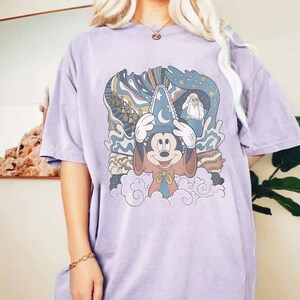 Puede incluir: Camiseta lila claro con un dise&ntilde;o de Mickey Mouse como mago, con un sombrero y capa azules. El dise&ntilde;o incluye elementos arremolinados azules y blancos y una representaci&oacute;n de Merl&iacute;n. La camiseta tiene un ajuste holgado y c&oacute;modo.