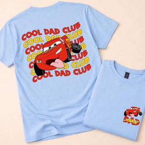 May include: Two light blue t-shirts. One features a large graphic of a red race car with the text "COOL DAD CLUB" in yellow and red. The other shirt has a smaller car graphic with the word "DAD".