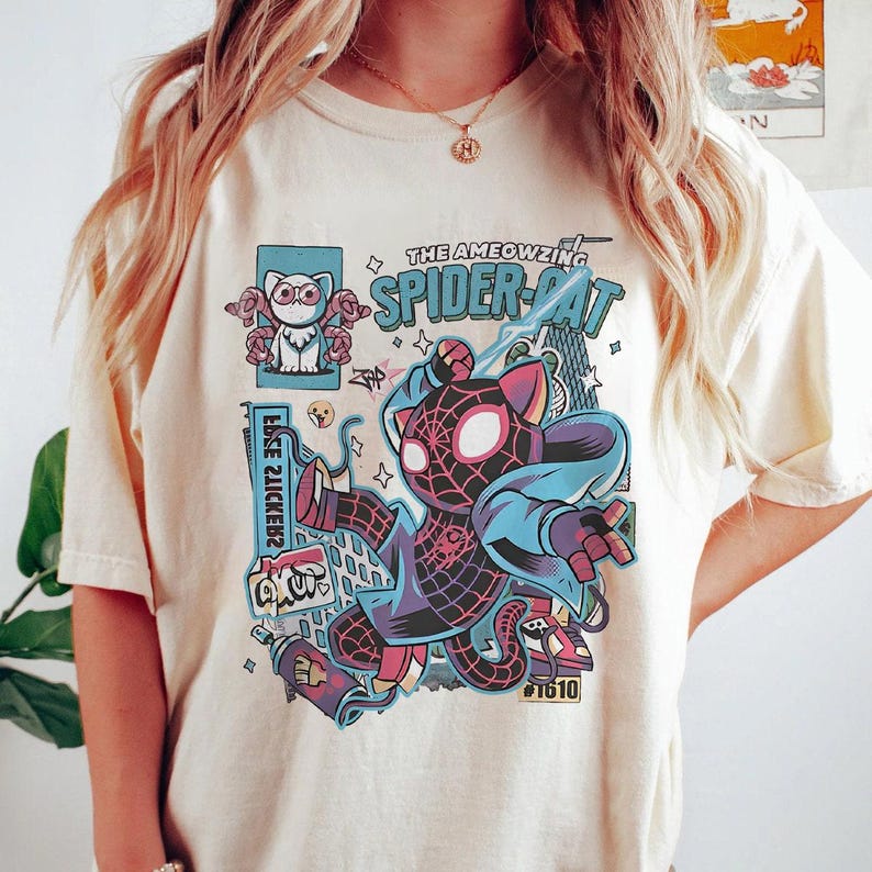 Puede incluir: Camiseta color crema con un gr&aacute;fico colorido de un gato con un disfraz inspirado en Spider-Man. El dise&ntilde;o incluye el texto "THE AMEOWZING SPIDER-CAT" y otros elementos de dibujos animados. Es una prenda informal.