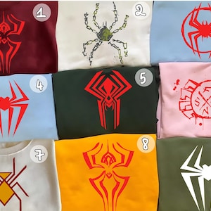 May include: A collection of nine sweatshirts, each featuring a unique spider-themed graphic in various colors. Designs include stylized spider emblems and web patterns in red, white, and yellow on maroon, white, blue, pink, green, and yellow backgrounds.