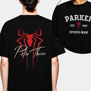 May include: Black t-shirt featuring a red spider-like emblem and the words "Peter Three" in white script. A smaller image shows the front of the shirt with "Parker" above "Spider-Man" and the year "1983".