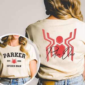 May include: Cream-colored t-shirt featuring a red and black Spider-Man design. The front of the shirt displays "PARKER EST. 1996 SPIDER-MAN" in black lettering. The back has a red spider emblem with "Peter One" in black script.