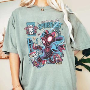 Puede incluir: Camiseta verde claro con una ilustraci&oacute;n de dibujos animados de un gato disfrazado de Spider-Man. El dise&ntilde;o incluye el texto "THE AMEOWZING SPIDER-CAT" y otros elementos gr&aacute;ficos. Prenda de vestir informal.