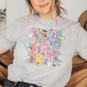 May include: A light gray t-shirt with the text "WHERE DREAMS COME TRUE" in colorful, retro-style lettering. The design includes cartoon characters, flowers, and stars. The shirt is short-sleeved.
