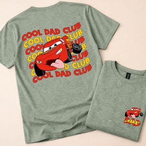 May include: Two heather green t-shirts. One features a red cartoon race car graphic with the text "COOL DAD CLUB" in yellow and red. The other shirt has a smaller car graphic with the word "DAD".