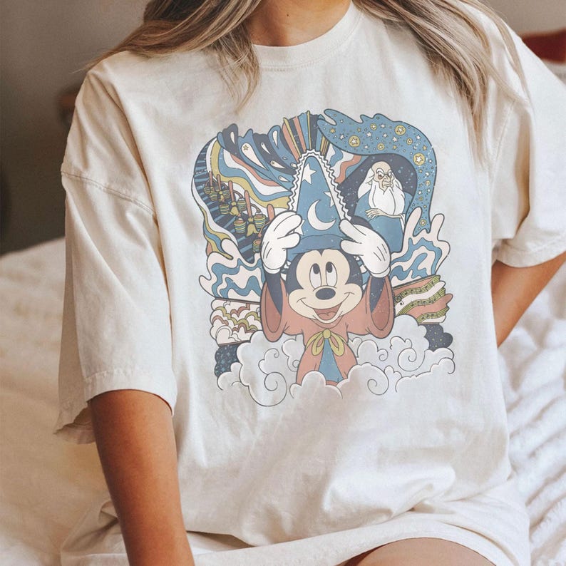Puede incluir: Camiseta color crema con un gr&aacute;fico colorido de Mickey Mouse con un sombrero de mago, rodeado de dise&ntilde;os azules y arco&iacute;ris. El dise&ntilde;o incluye un personaje con una larga barba blanca. La camiseta tiene mangas cortas.