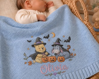 Könnte beinhalten: Eine hellblaue Strickdecke für Babys mit einer Stickerei von Winnie Puuh und I-Aah, die Hexenhüte tragen, umgeben von Kürbissen, Fledermäusen und Sternen. Der Name Olivia ist darunter in rosafarbener Schreibschrift eingestickt.