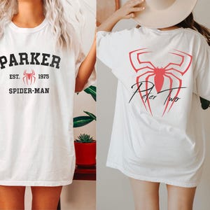 May include: White t-shirt featuring the text "PARKER EST. 1975 SPIDER-MAN" in black and red. The back of the shirt displays a red spider graphic with the text "Peter Two" in black script. The shirt is a casual, oversized fit.
