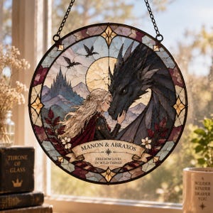 Könnte beinhalten: Ein runder Buntglas-Suncatcher, der eine blonde Person zeigt, die mit der Stirn einen großen schwarzen Drachen berührt. Das Stück enthält ein Banner mit dem Text MANON & ABRAXOS und FREEDOM LIVES IN WILD THINGS vor einer Berglandschaft.