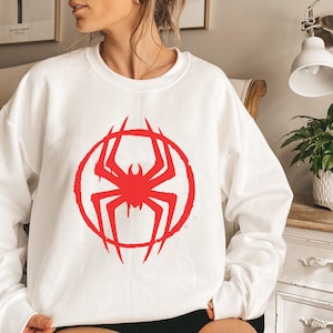 May include: White crewneck sweatshirt featuring a red spider graphic inside a red circle. The spider design has a distressed, graffiti-style aesthetic. The sweatshirt is a casual, long-sleeved garment.