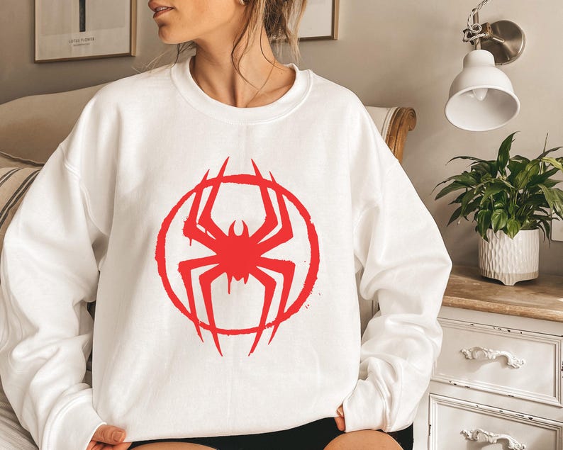 May include: White crewneck sweatshirt featuring a red spider graphic inside a red circle. The spider design has a distressed, graffiti-style aesthetic. The sweatshirt is a casual, long-sleeved garment.