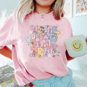 May include: Pink crew neck t-shirt with the text "WHERE DREAMS COME TRUE" in a colorful, retro font. The design includes cartoon characters and floral accents. A white mug with a smiley face is held in the hand.
