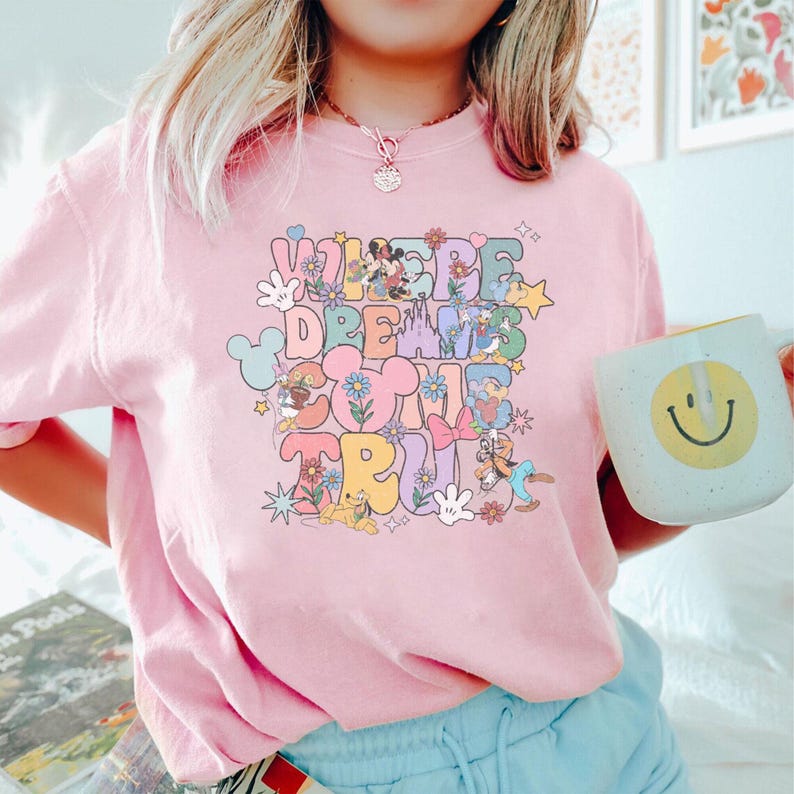 May include: Pink crew neck t-shirt with the text "WHERE DREAMS COME TRUE" in a colorful, retro font. The design includes cartoon characters and floral accents. A white mug with a smiley face is held in the hand.