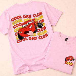 May include: Two light pink t-shirts. One shirt features a large graphic of a red race car with the text "COOL DAD CLUB" in yellow and red. The other shirt has a smaller version of the car graphic with the word "DAD".