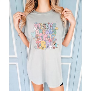 May include: A light green t-shirt with the text "WHERE DREAMS COME TRUE" in a colorful, whimsical design. The design includes cartoon characters, flowers, and stars. The shirt is a relaxed fit.
