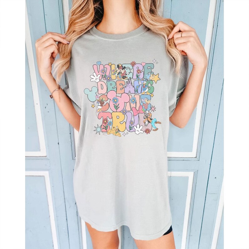 May include: A light green t-shirt with the text "WHERE DREAMS COME TRUE" in a colorful, whimsical design. The design includes cartoon characters, flowers, and stars. The shirt is a relaxed fit.