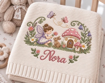 May include: A cream-colored knit baby blanket featuring an embroidered scene of a fairy, a rabbit, and a mushroom house surrounded by flowers and butterflies. The name Nora is embroidered in pink script below the scene, with small floral accents.