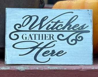 May include: A rectangular wooden sign with a distressed white finish and black lettering that reads Witches GATHER Here. The text features a mix of cursive and block fonts with decorative flourishes, presenting a weathered appearance on the surface.