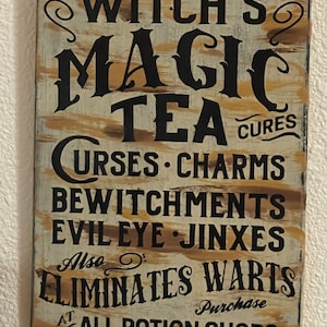May include: A rectangular wooden sign with a distressed beige and brown painted background featuring black text that reads: WITCH'S MAGIC TEA CURES CURSES CHARMS BEWITCHMENTS EVIL EYE JINXES ALSO ELIMINATES WARTS PURCHASE AT ALL POTION SHOPS.