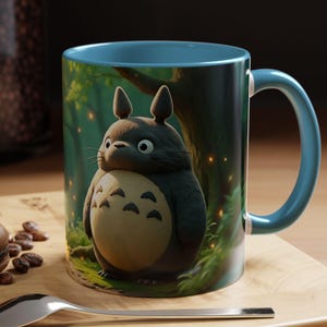 Könnte beinhalten: Eine Keramik-Tasse mit blauem Griff und Innenraum zeigt eine skurrile Illustration eines Totoro-Charakters in einer Waldszene. Die Tasse steht auf einer Holzoberfläche mit einem Löffel und einem Macaron.