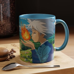 Op de afbeelding: Een blauwe keramische mok met een kleurrijke anime-illustratie van een personage dat op een vlam blaast. De mok heeft een blauwe handgreep en binnenkant, met een landschapsachtergrond. Een lepel en een macaron staan op de voorgrond.
