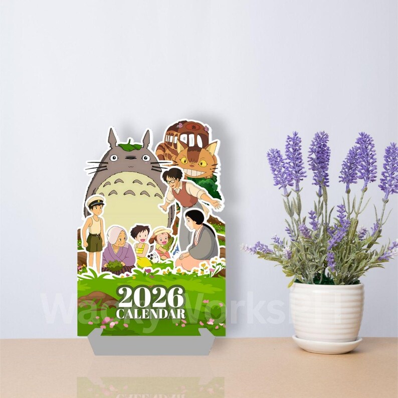 May include: A 2026 calendar featuring characters from the animated film My Neighbor Totoro. The calendar has a green and white base with a colorful illustration of Totoro and other characters. The calendar is displayed on a wooden surface next to a potted lavender plant.